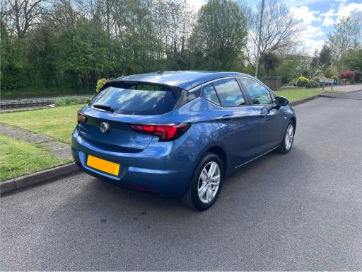 Blue hatchback car parked on a residential street, surrounded by greenery.