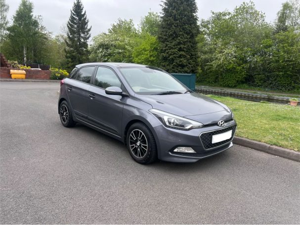 A grey hatchback car parked on a road with greenery in the background.