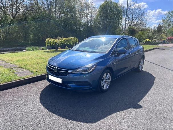 Blue hatchback car parked on a sunny road with greenery in the background.